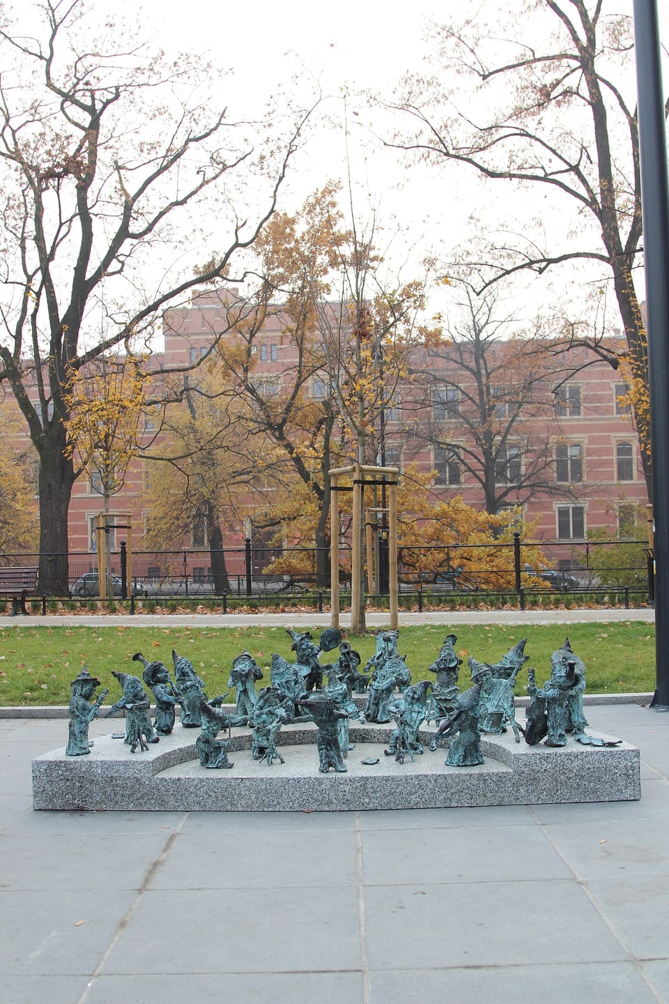 Eine Gruppe kleiner Zwergfiguren aus Bronze mit Instrumenten, wie ein Orchester auf einem geschwungenen Granitsockel auf einem gepflasterten Platz aufgestellt, dahinter kahle Bäume und ein Backsteingebäude.