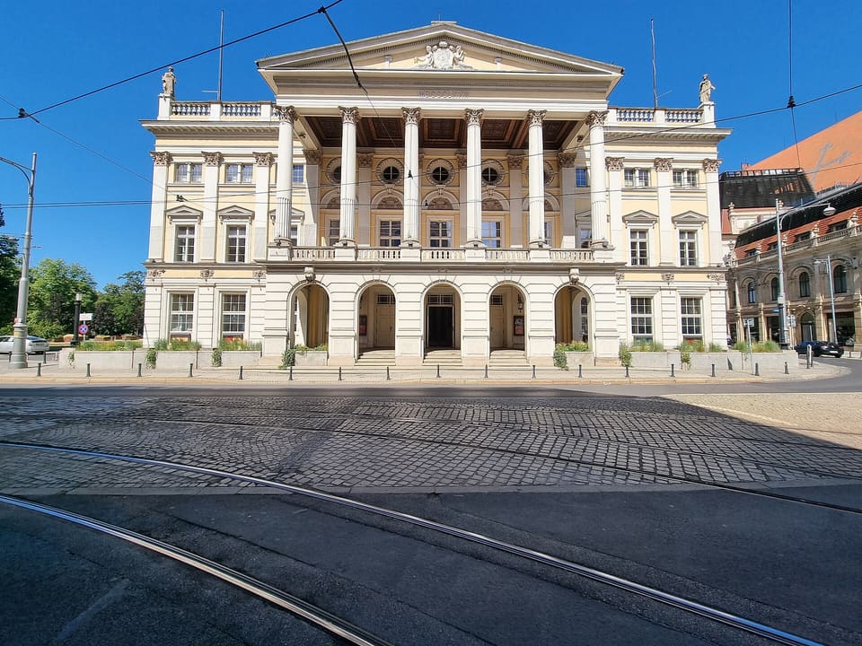 Frontalansicht des creme-gelben klassizistischen Opernhauses mit mittigem Säulenportikus und Giebel unter klarem blauem Himmel; Straßenbahngleise queren die leere Straße davor.