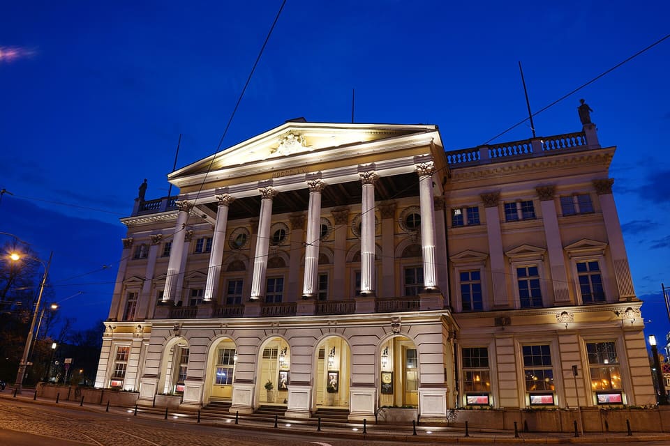 The opera house lit after dark, its columned portico and facade illuminated against a deep blue sky, seen from the street corner.