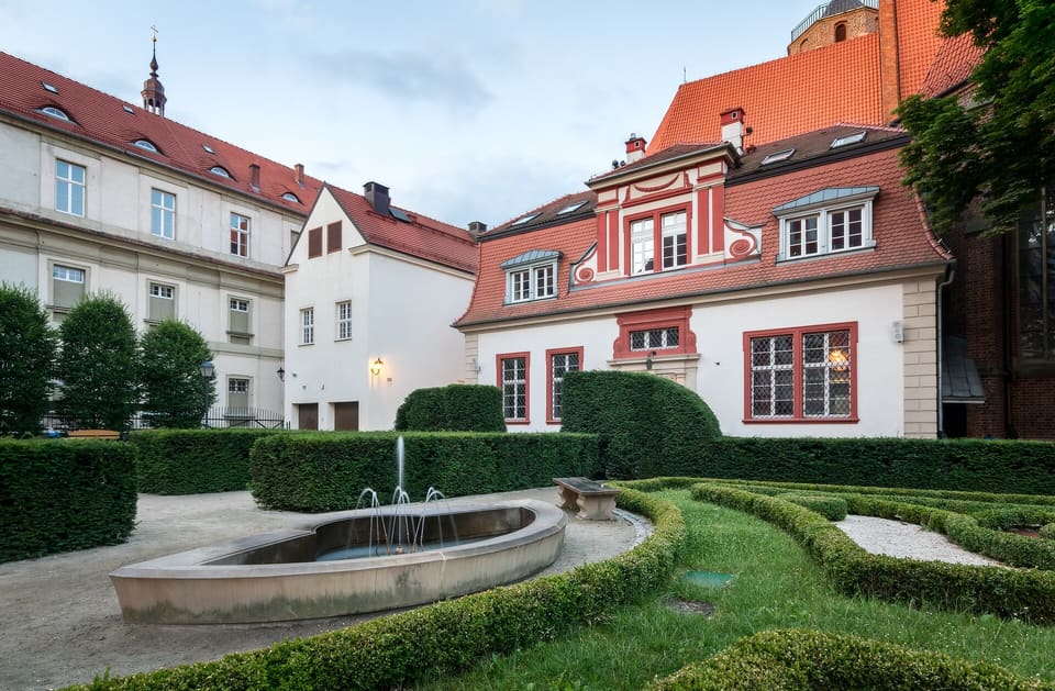 The baroque courtyard garden in the evening, with a stone fountain, clipped box hedges and the building's red-trimmed facade behind.