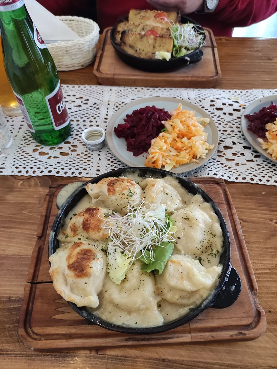 Eine gusseiserne Pfanne mit gebratenen Pierogi in Sahnesoße, garniert mit Sprossen und Salat, auf einem Holzbrett, daneben Teller mit Rotkohl- und Karottensalat und eine grüne Flasche auf einer gehäkelten Tischdecke.