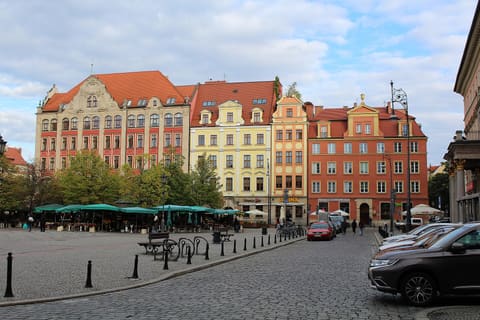 A wide paved square with green-roofed market stalls in the middle, a row of red, yellow and cream tenement houses behind them, and a small tour vehicle on the right.
