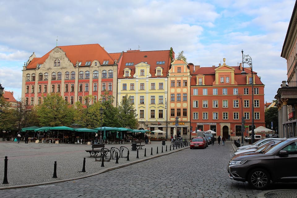 A wide paved square with green-roofed market stalls in the middle, a row of red, yellow and cream tenement houses behind them, and a small tour vehicle on the right.