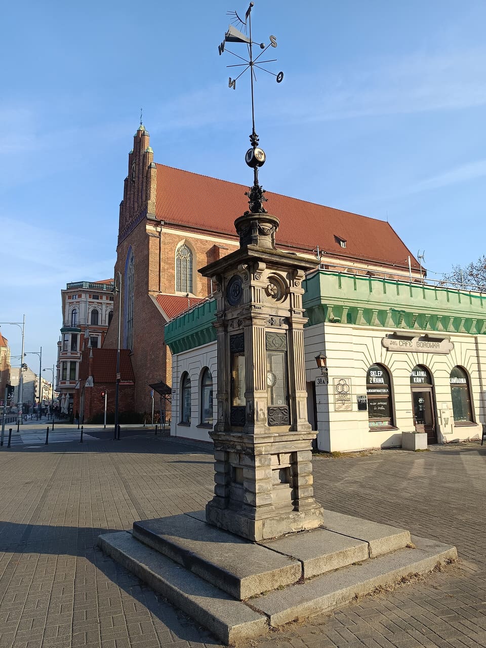 Die kleine meteorologische Säule aus Sandstein auf ihrem gestuften Sockel, mit leeren Instrumentennischen und einem eisernen Mast darüber, der eine Wetterfahne und eine Windrose mit den Buchstaben N, S, O und W trägt.
