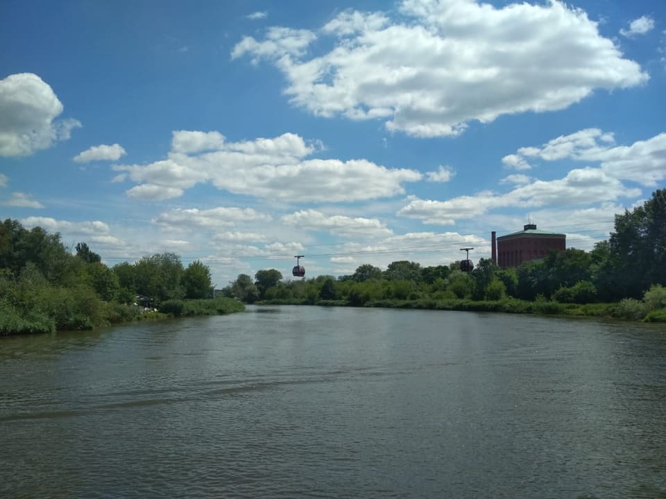 Zwei rote Gondeln hängen an Seilen über der Oder, vom Ufer aus gesehen, mit einem Wasserturm aus Backstein und Bäumen auf der anderen Seite unter blauem Himmel.