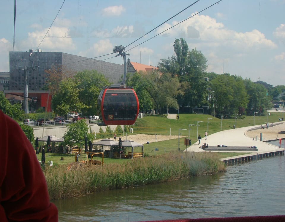 Eine rote Gondel hängt an Seilen über einem grasbewachsenen Ufer mit Bäumen, einem Fußweg und einem Universitätsgebäude, gesehen über das Wasser der Oder.