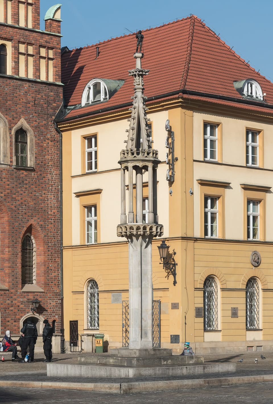 The Gothic pillory column topped by a small figure stands on its stepped base in front of a yellow corner house and the brick wall of the Old Town Hall.