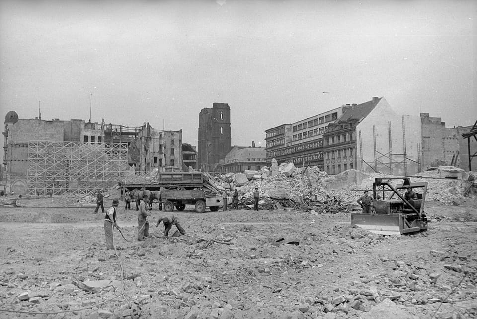 Workers clearing rubble by hand beside a truck and a bulldozer on a cleared site, with gutted buildings and a damaged tower standing behind.
