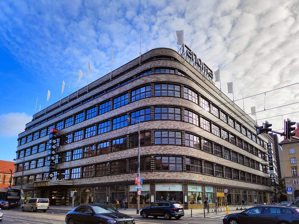 Die abgerundete Ecke des Kaufhauses Renoma mit waagerechten Fenster- und Backsteinbändern, einem Schriftzug auf dem Dach und Fahnen, von der Straße aus gesehen unter blauem Himmel mit Wolken.