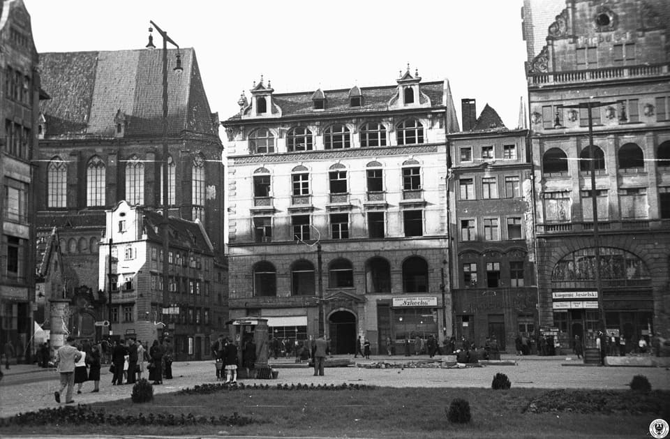 Damaged tenement houses on the Market Square in 1947, their upper floors burnt out, with people walking past and a gutted building at the corner.