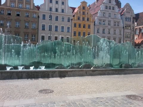 A low fountain of upright glass panes with water running down them and jets rising in front, with the tenement houses of the square behind.