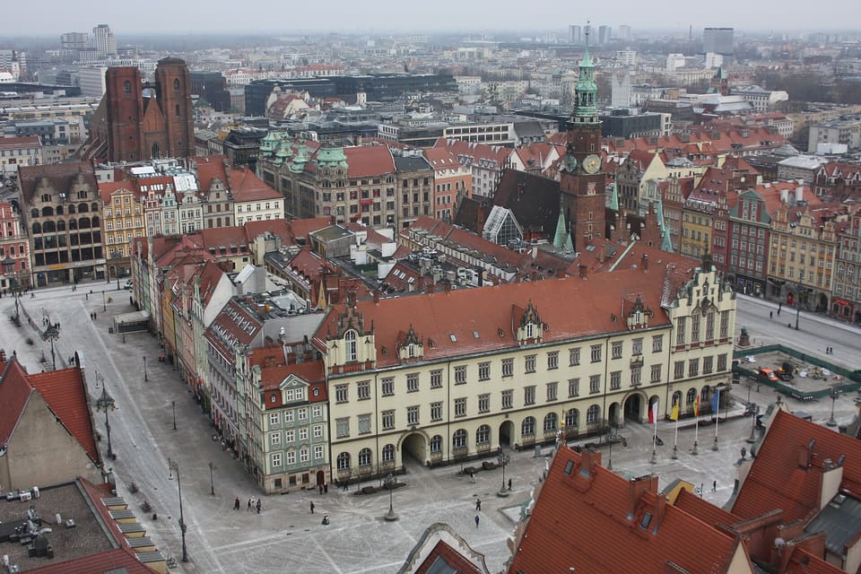 A view down onto the Rynek from a church tower, showing the Old Town Hall's tower and gabled roofline, rows of houses lining the square, and brick church towers in the distance.