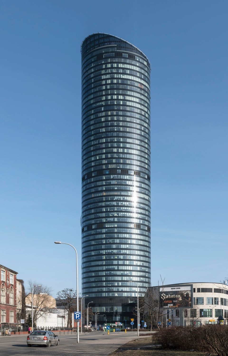 Der zylindrische gläserne Sky Tower von einer Straße unten aus gesehen, unter klarem blauem Himmel, mit älteren Backsteinhäusern und einem geparkten Auto an seinem Fuß.