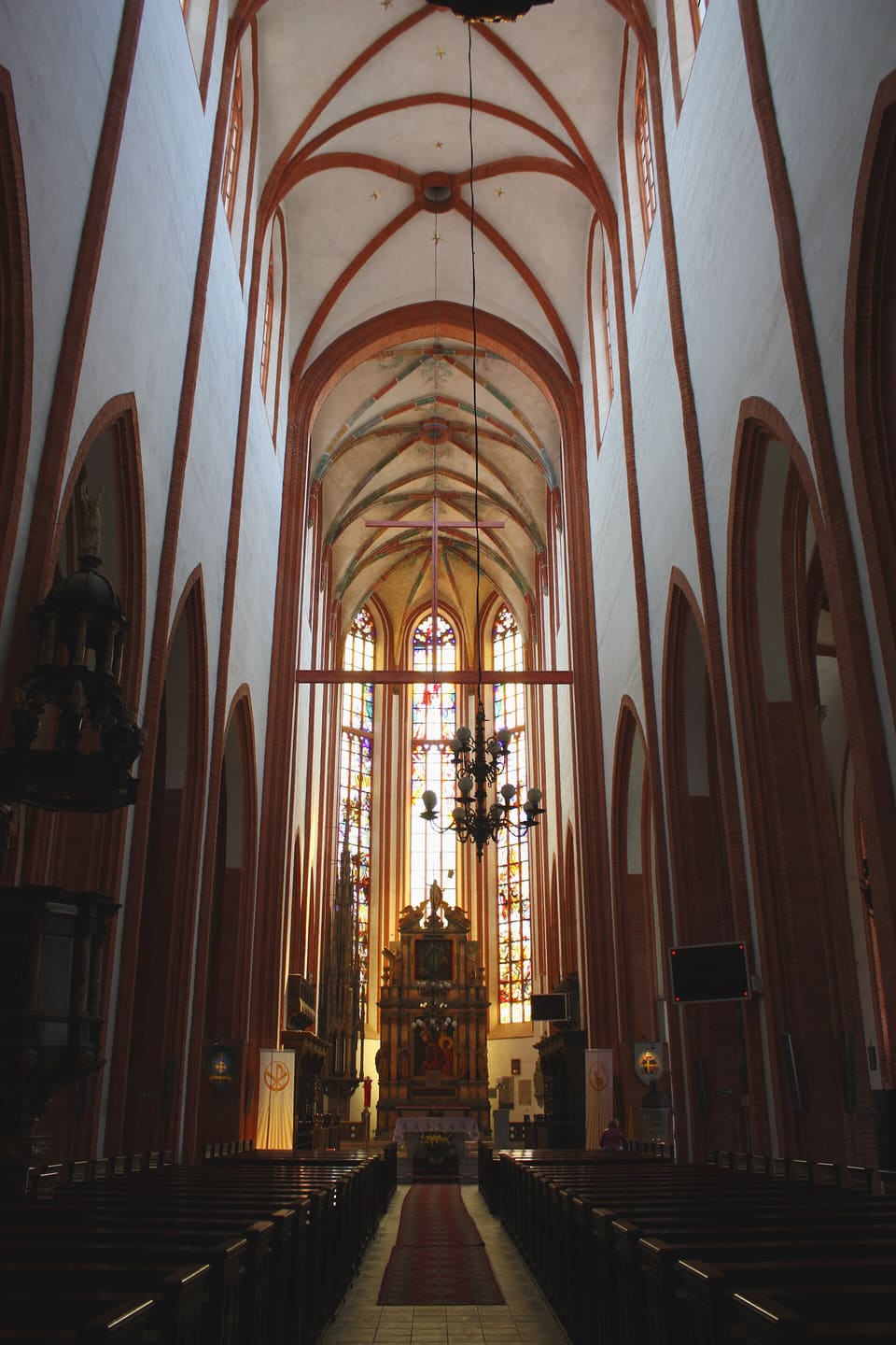 Blick durch das Hauptschiff von St. Elisabeth zum Altar, mit gotischem Rippengewölbe darüber, hohen Fenstern, einem schmiedeeisernen Kronleuchter und Reihen leerer Kirchenbänke.