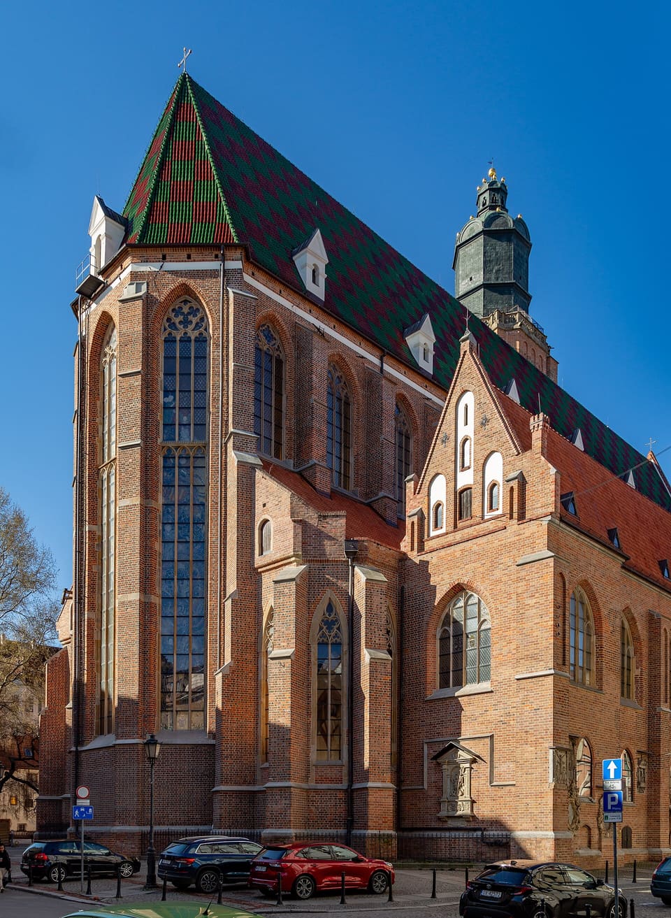 Die gotische Backsteinkirche von Nordosten, mit einem hohen, grün und rot gemusterten Ziegeldach, dahinter ein dunkler Turm mit Kupferhaube und ein klarer blauer Himmel.