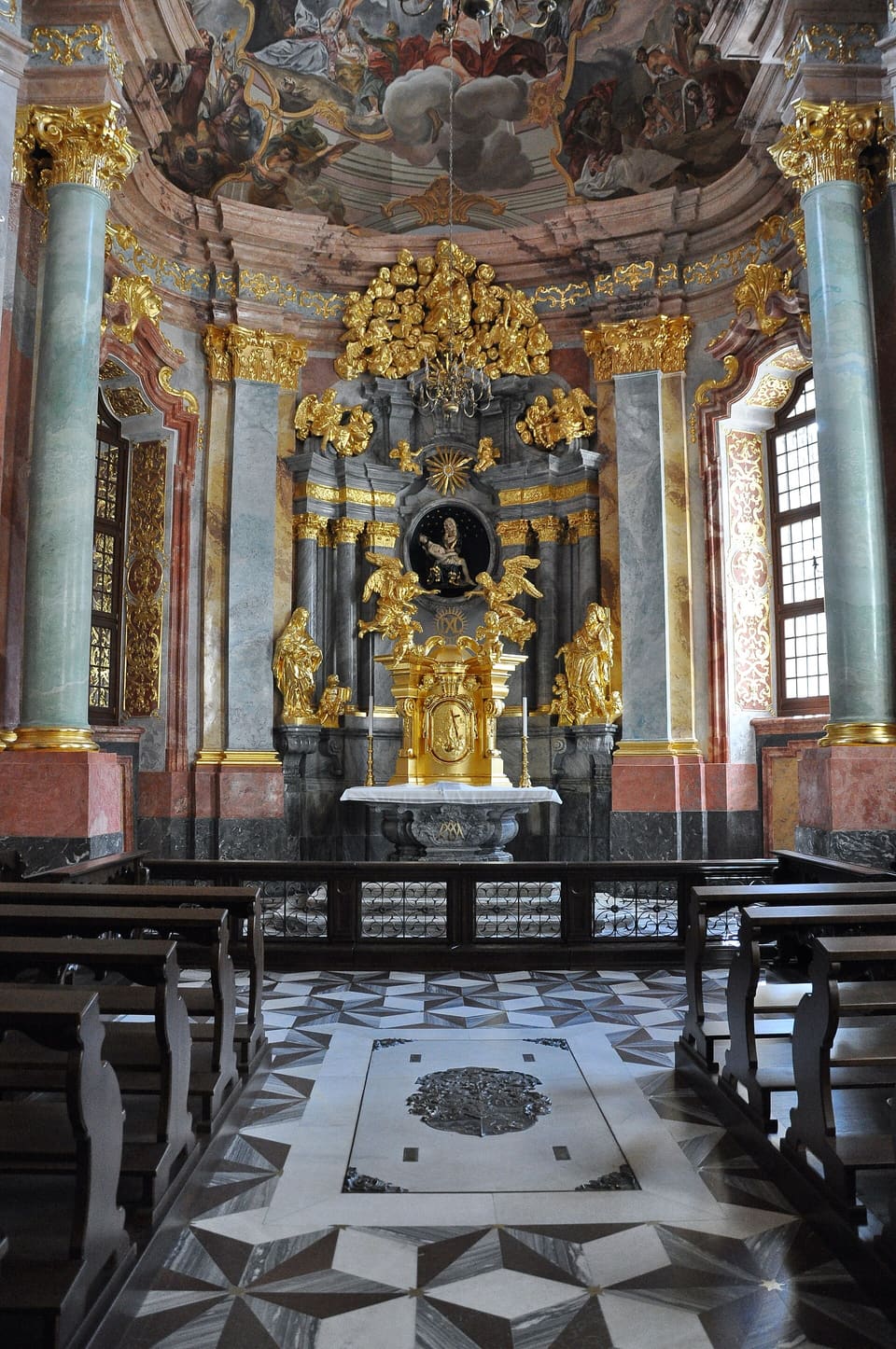 Inside the Hochberg Chapel: a gilded Baroque altar between grey-green marbled columns, a painted dome above, and a black-and-white patterned stone floor between dark pews.