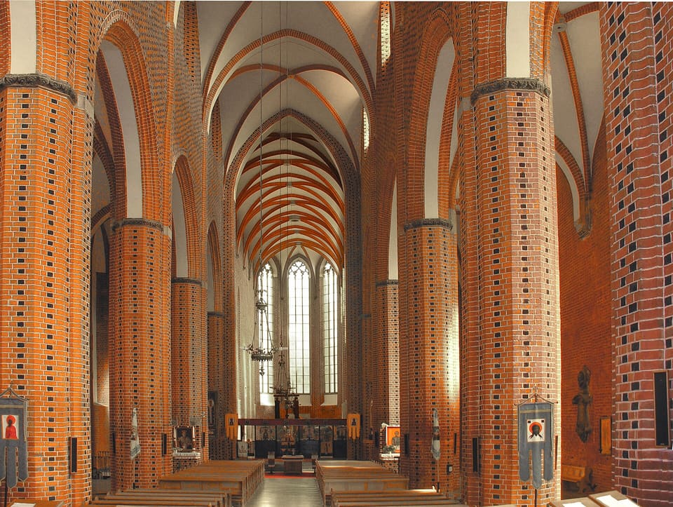 Looking east down the nave between round brick pillars to three tall lancet windows, under red-ribbed Gothic vaults, with rows of wooden pews and icons hung on the pillars.