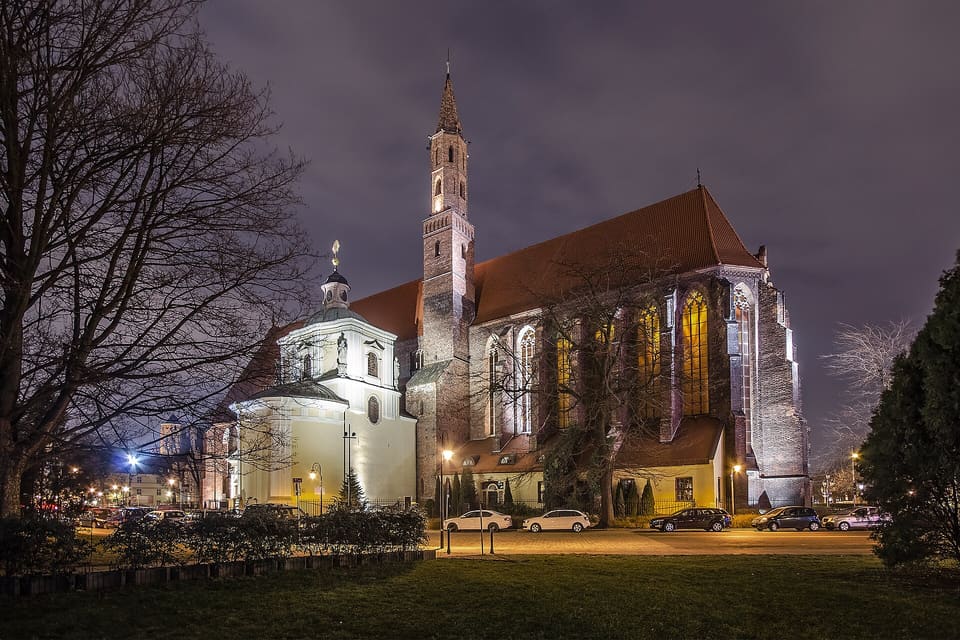 The red-brick Gothic church at night, its tall windows lit from inside, a slender brick tower in the middle and the cream Baroque Hochberg Chapel with its small domed lantern at the left, seen across a lawn and a row of parked cars.