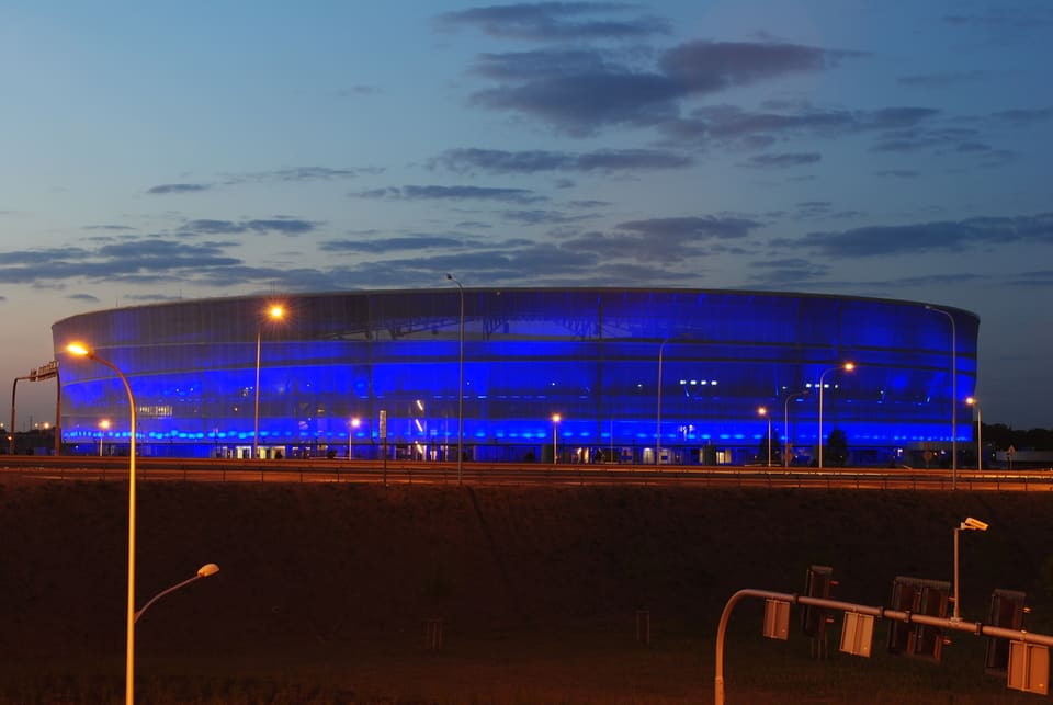 Die durchscheinende Fassadenverkleidung des ovalen Stadions leuchtet nachts tiefblau, gesehen von der Zufahrtsstraße mit Straßenlaternen und den Lichtspuren vorbeifahrender Autos.