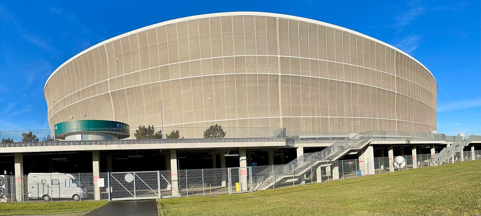 Das ovale Stadion mit seiner hellen Fassadenverkleidung von außen unter blauem Himmel, mit einem Grashang und einem am Fuß geparkten Transporter.