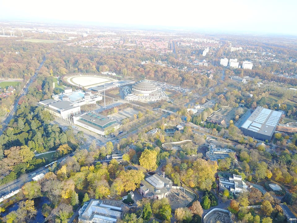 Die Kuppel der Hala Stulecia, vom gepflasterten Platz davor gesehen, mit einem Säuleneingang, Reihen von Rundbogenfenstern und einigen Besuchern, die den Platz unter klarem Himmel überqueren.