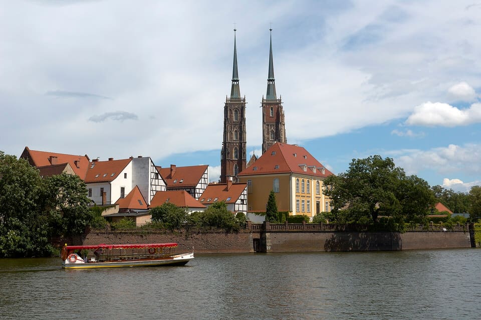 Ein kleines weißes Passagierboot mit rotem Sonnendach auf der Oder, unterhalb der Ziegelufermauer des Ostrów Tumski, dahinter ragen die zwei Turmspitzen der Kathedrale über rot gedeckten Häusern und Fachwerkhäusern auf.