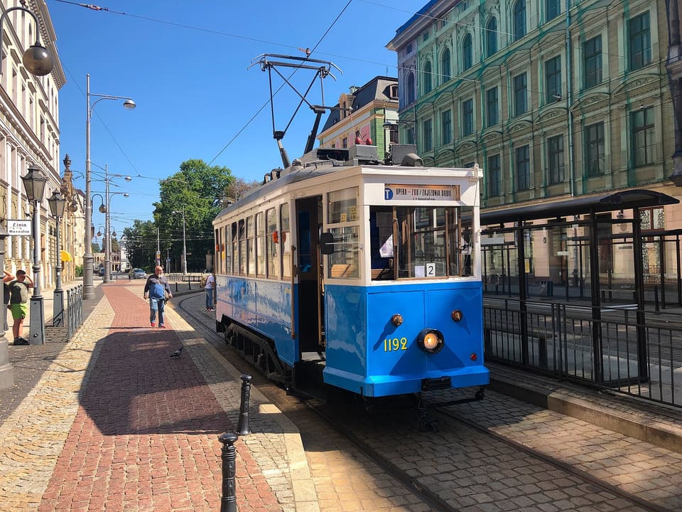 Eine blau-cremefarbene zweiachsige Straßenbahn mit der Nummer 1192, mit einem Stromabnehmer auf dem Dach und einem Zielschild, das die Linie T zum Zoo zeigt, an einem sonnigen Tag an einer Haltestelle in einer Kopfsteinpflasterstraße, dahinter hohe Häuser aus dem neunzehnten Jahrhundert.