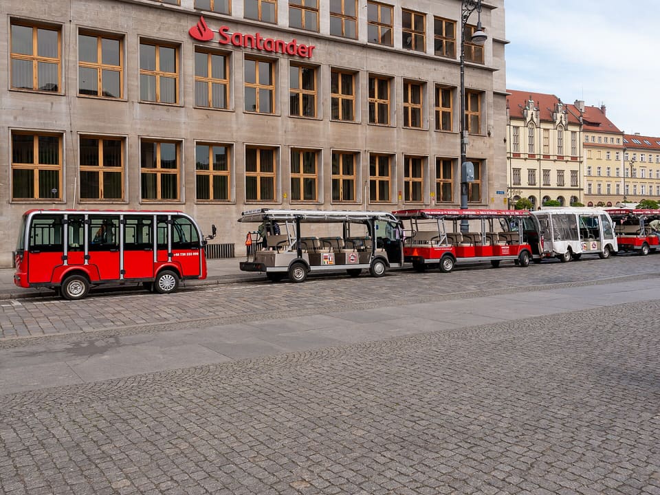 Eine Reihe kleiner elektrischer Besichtigungswagen, am Rand des Marktplatzes geparkt: vorn ein geschlossener roter mit verglasten Türen, dann offene Wagen mit Sonnendächern und Sitzbankreihen, vor einem steinernen Bürogebäude, am anderen Ende Giebelhäuser.