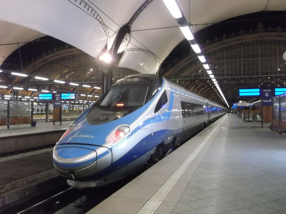 A silver and blue Pendolino high-speed train standing at a platform of Wrocław Główny under the white membrane roof, with the old iron and glass platform halls behind it.