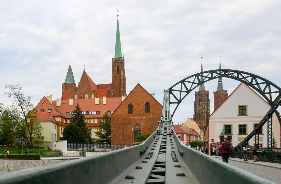 Looking east along the riveted green girder of Tumski Bridge: the Holy Cross church with its single green spire on the left, and the cathedral's two spired towers under the bridge's arch on the right.