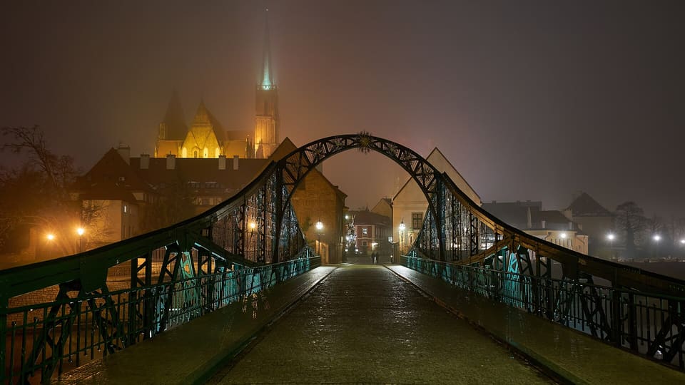The green iron Tumski Bridge at night, seen from the bridge itself toward Ostrów Tumski, its lamps lit and the illuminated spires of the cathedral beyond.