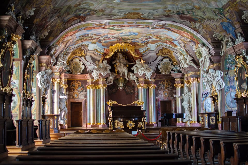 Die Aula Leopoldina von der Rückseite des Saals: unten dunkle Holzbänke, an den Wänden marmorierte Säulen und weiße Stuckfiguren, ein vergoldeter Baldachin über dem Rednerpult und das ganze Tonnengewölbe mit einem Fresko aus Figuren und Himmel bemalt.