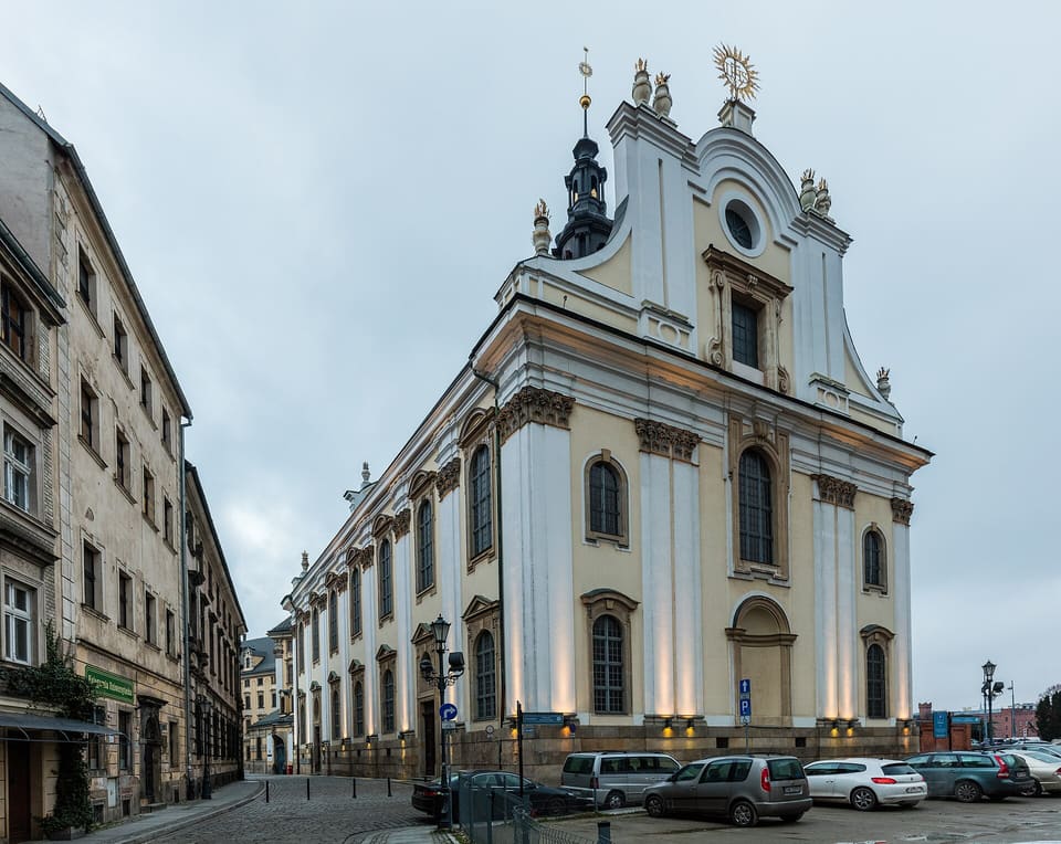 The University Church seen from the street: a long cream and white baroque wall with tall arched windows, ending in a curved gable crowned by a gilded IHS sunburst.