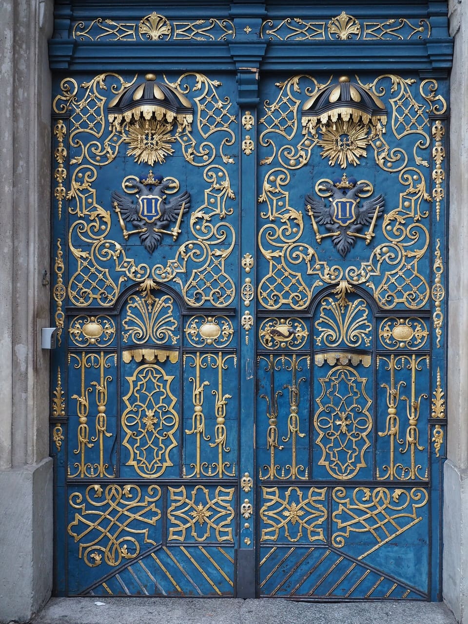 Eine hohe, blau gestrichene Doppeltür mit goldfarbenen schmiedeeisernen Ranken, oben zwei gekrönte Embleme.