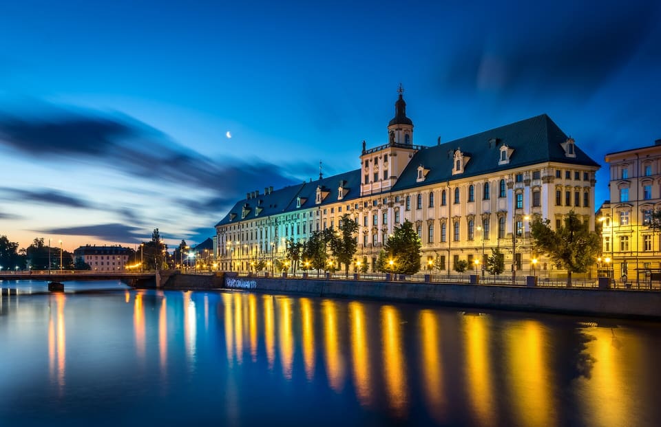 Das Hauptgebäude der Universität an der Oder unter tiefblauem Himmel, die Fenster erleuchtet und die Lichter im Fluss gespiegelt.