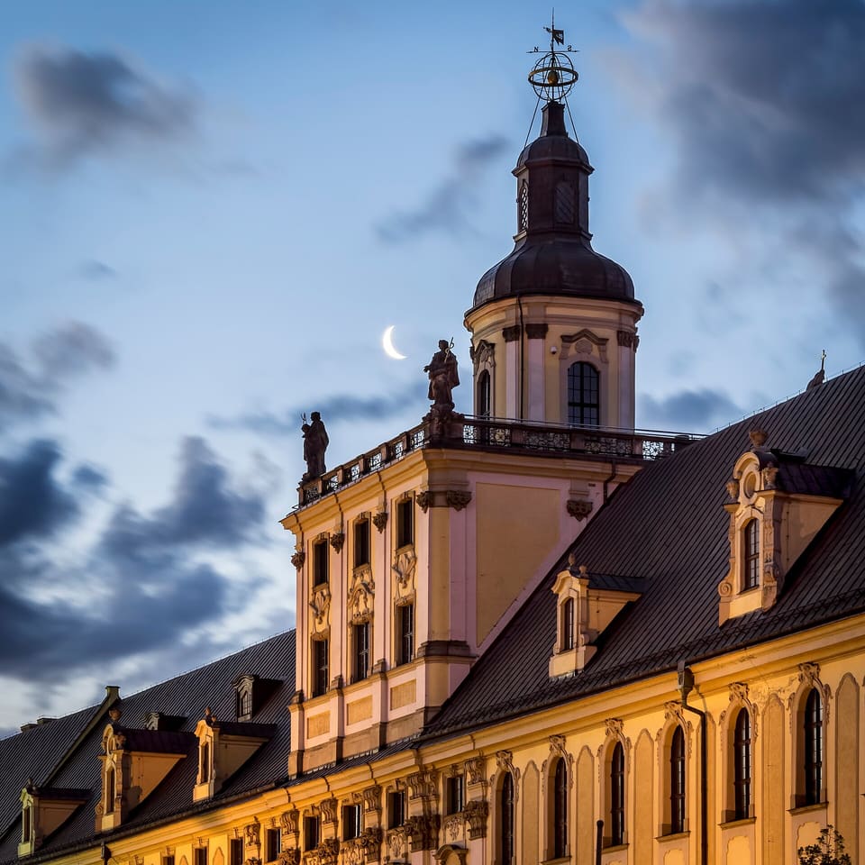 Der Mathematische Turm in der Abenddämmerung beleuchtet über der Dachlinie der Universität, Kuppel und vergoldete Spitze vor tiefblauem Himmel, zwei der allegorischen Figuren auf der Terrassenbalustrade als Silhouette, eine Mondsichel neben dem Turm.