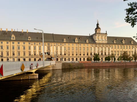 The long yellow facade of the university's main building beside the Odra, with the Mathematical Tower rising from its centre, seen from the bridge.