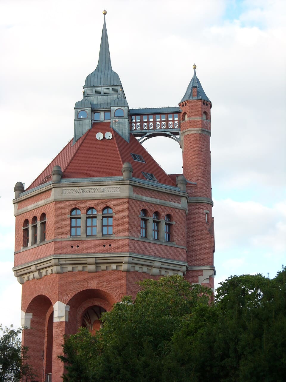 Nahaufnahme der Spitze des Backstein-Wasserturms mit seiner kegelförmigen Turmspitze, dem runden Ecktürmchen unter seinem eigenen spitzen Dach und der kleinen überdachten Brücke, die beide verbindet, vor blauem Himmel.