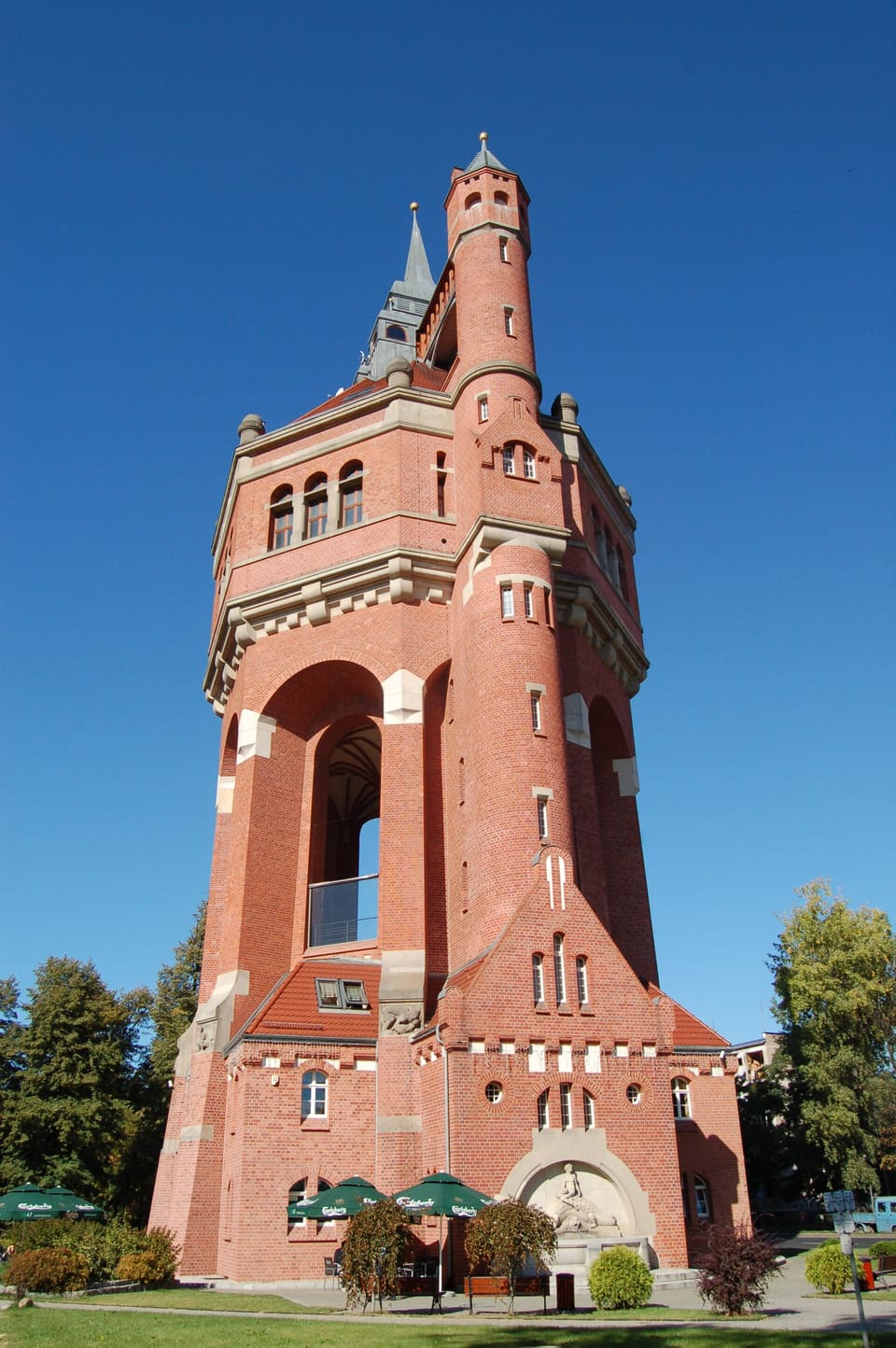 Der hohe Wasserturm aus rotem Backstein mit einem runden Ecktürmchen und einer Turmspitze, von seinem Fuß aus vor tiefblauem Himmel gesehen, mit Bäumen und grünem Rasen ringsum.