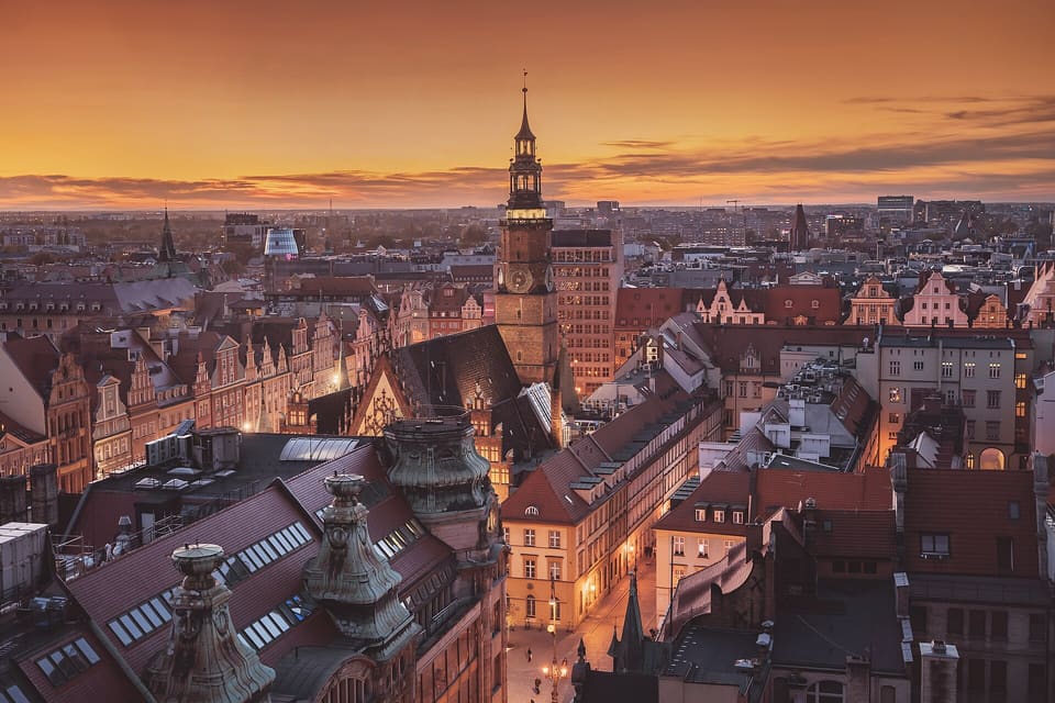 Looking west over the Old Town roofs at dusk from high up: the Town Hall's clock tower and its traceried gable rising above them, a lit street below, and an orange sky over the city beyond.