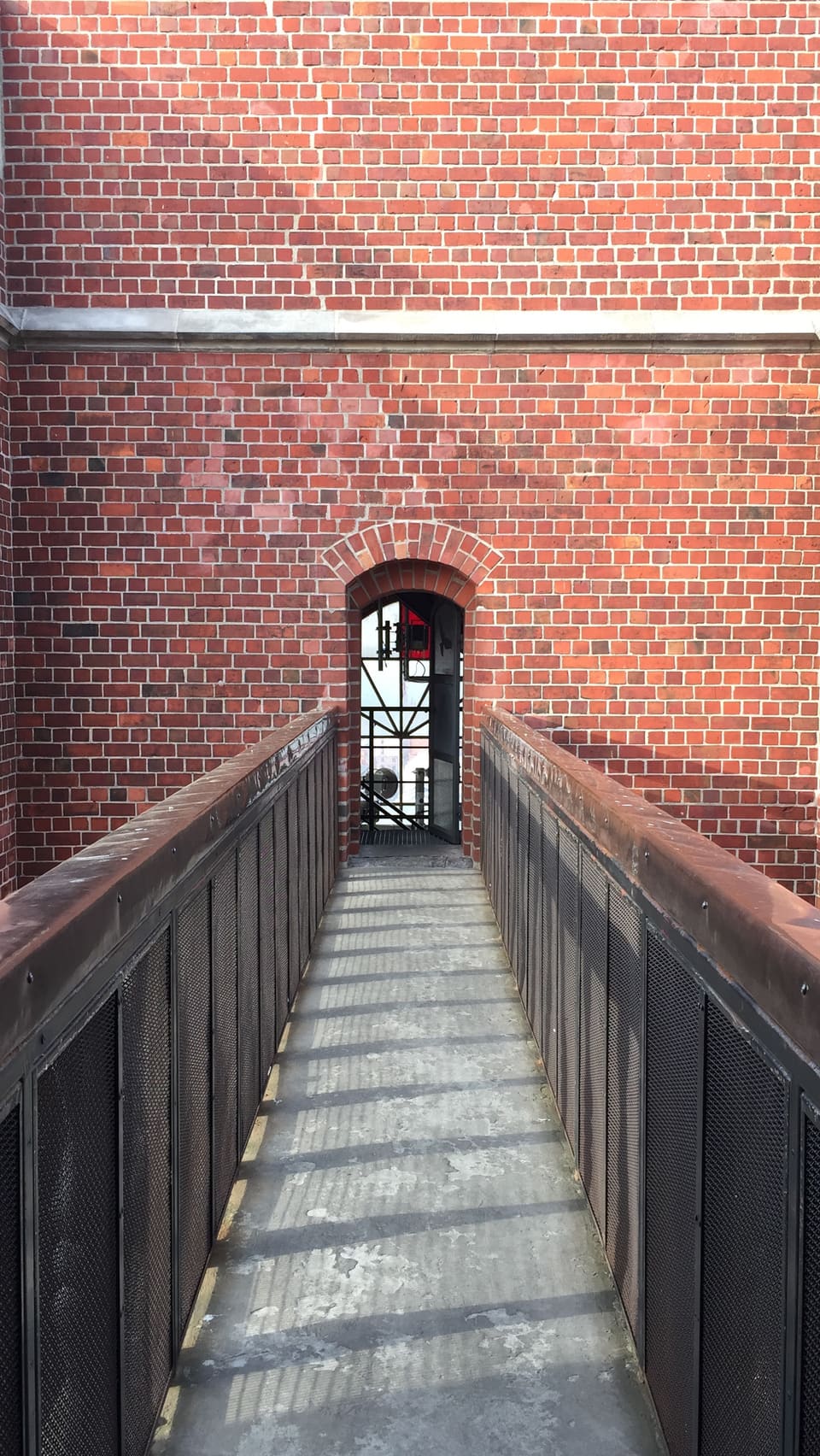 Auf der Brücke selbst stehend, der Blick entlang ihres schmalen Gangs zwischen dunklen Metallgeländern auf beiden Seiten zu einem kleinen Backsteinbogen voraus.