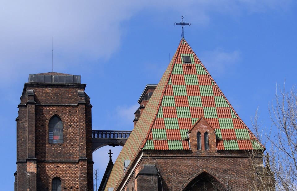 Ein dunkler Backstein-Kirchturm links, durch eine kurze Bogenbrücke mit Balustrade mit dem nächsten Turm verbunden, neben einem grün-rot karierten, ziegelgedeckten Giebeldach unter blauem Himmel.