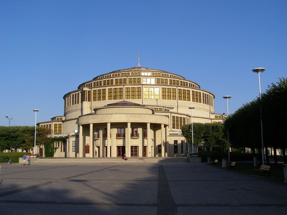 Der runde Kuppelbau der Hala Stulecia von außen im Licht des späten Nachmittags, davor ein gepflasterter Platz, Bänke, Bäume und einige kleine Gestalten.